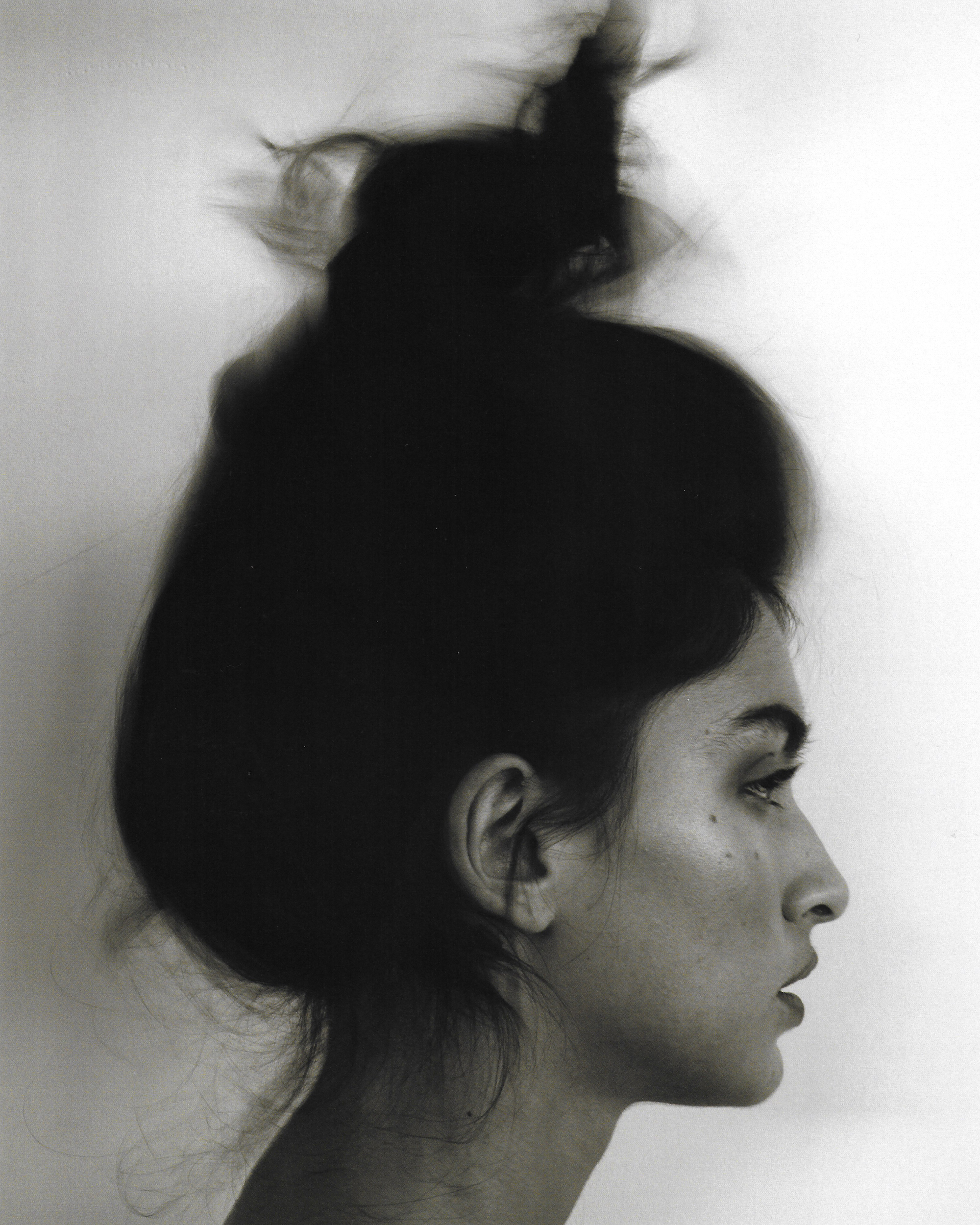 Profile portrait of a woman with dark hair in a high updo against a plain white background by Adriana Roslin
