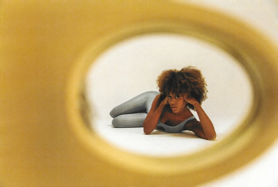 Portrait of a woman with curly brown hair lying on a white surface, framed by a large yellow circle, creating an artistic composition.