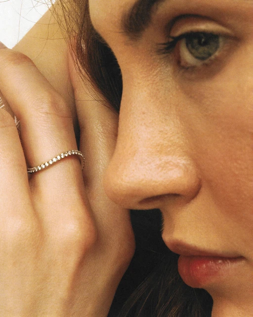 Close-up of a woman's face and hand, showcasing a diamond ring on her finger. The focus is on the ring and the woman's skin.