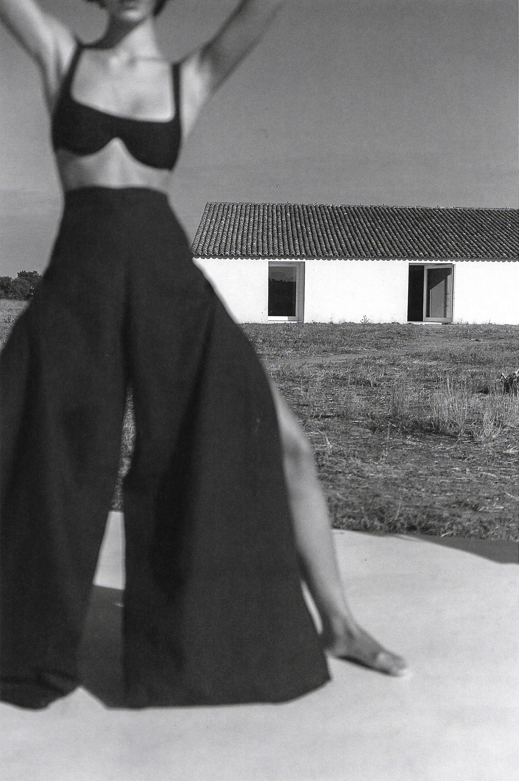 A monochrome fashion shot of a woman in a bikini top and wide-legged pants, raising her arms, posing in front of a white building.
