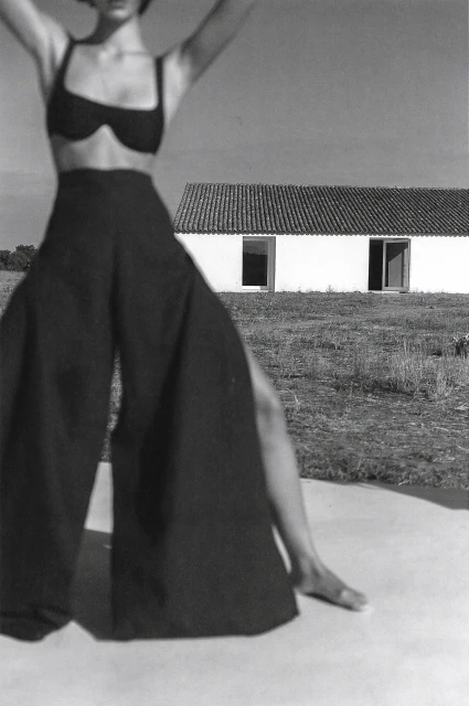A monochrome fashion shot of a woman in a bikini top and wide-legged pants, raising her arms, posing in front of a white building.