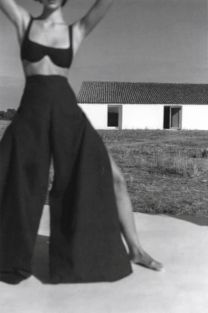 A monochrome fashion shot of a woman in a bikini top and wide-legged pants, raising her arms, posing in front of a white building.