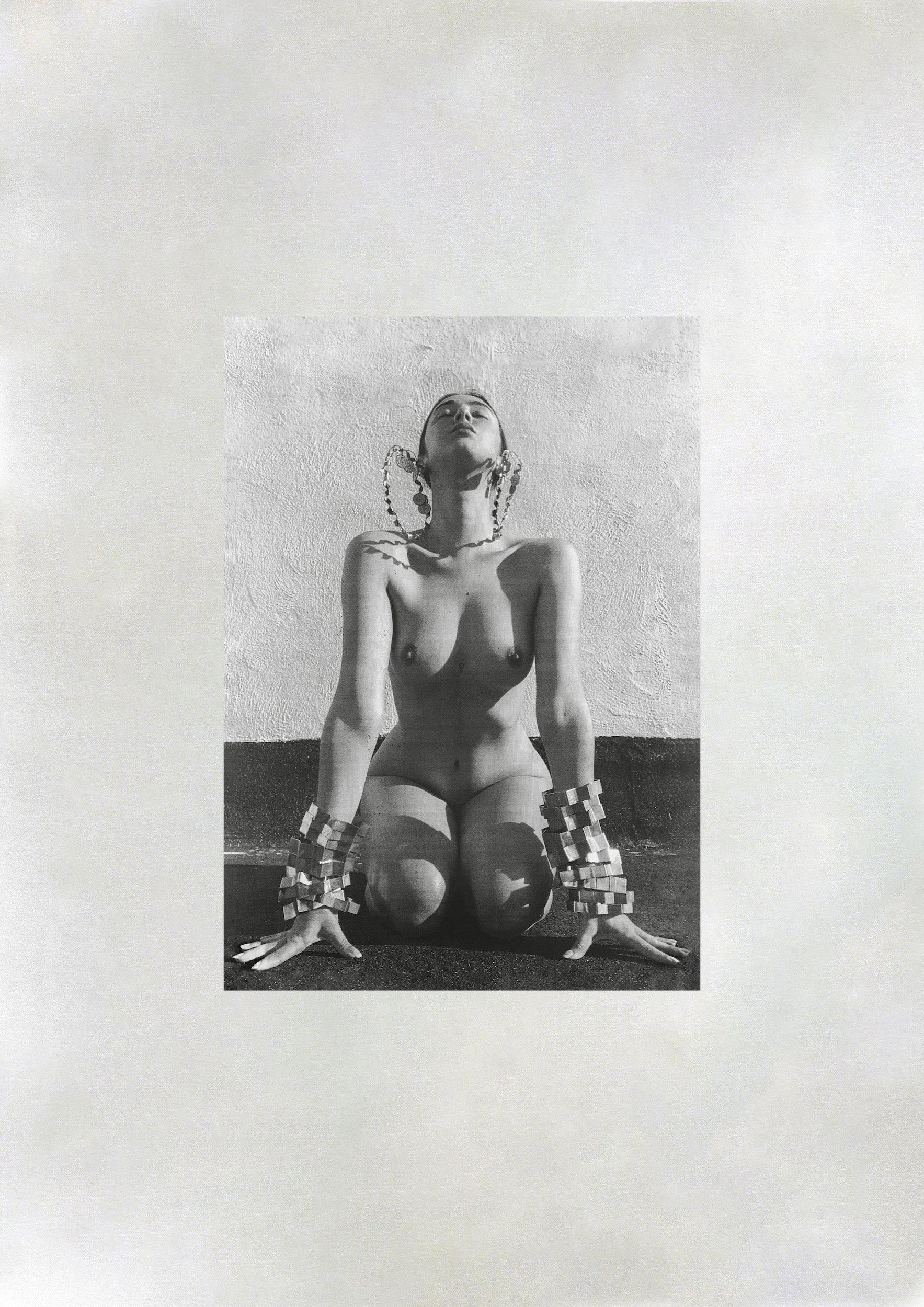 Monochrome photo of a nude woman kneeling with ornate bracelets and head jewelry, set against a lightly textured wall.