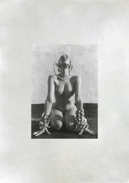 Monochrome photo of a nude woman kneeling with ornate bracelets and head jewelry, set against a lightly textured wall.