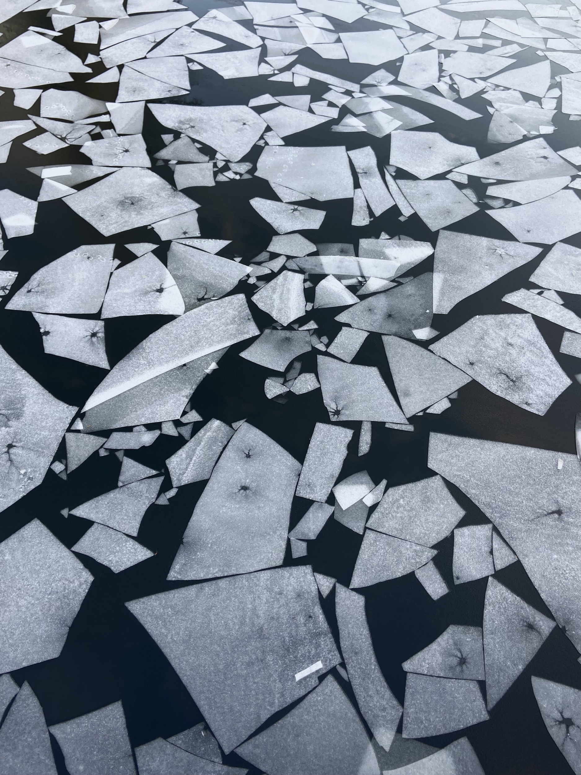 An aerial view of dark water with white ice fragments in a variety of shapes and sizes floating on the surface.