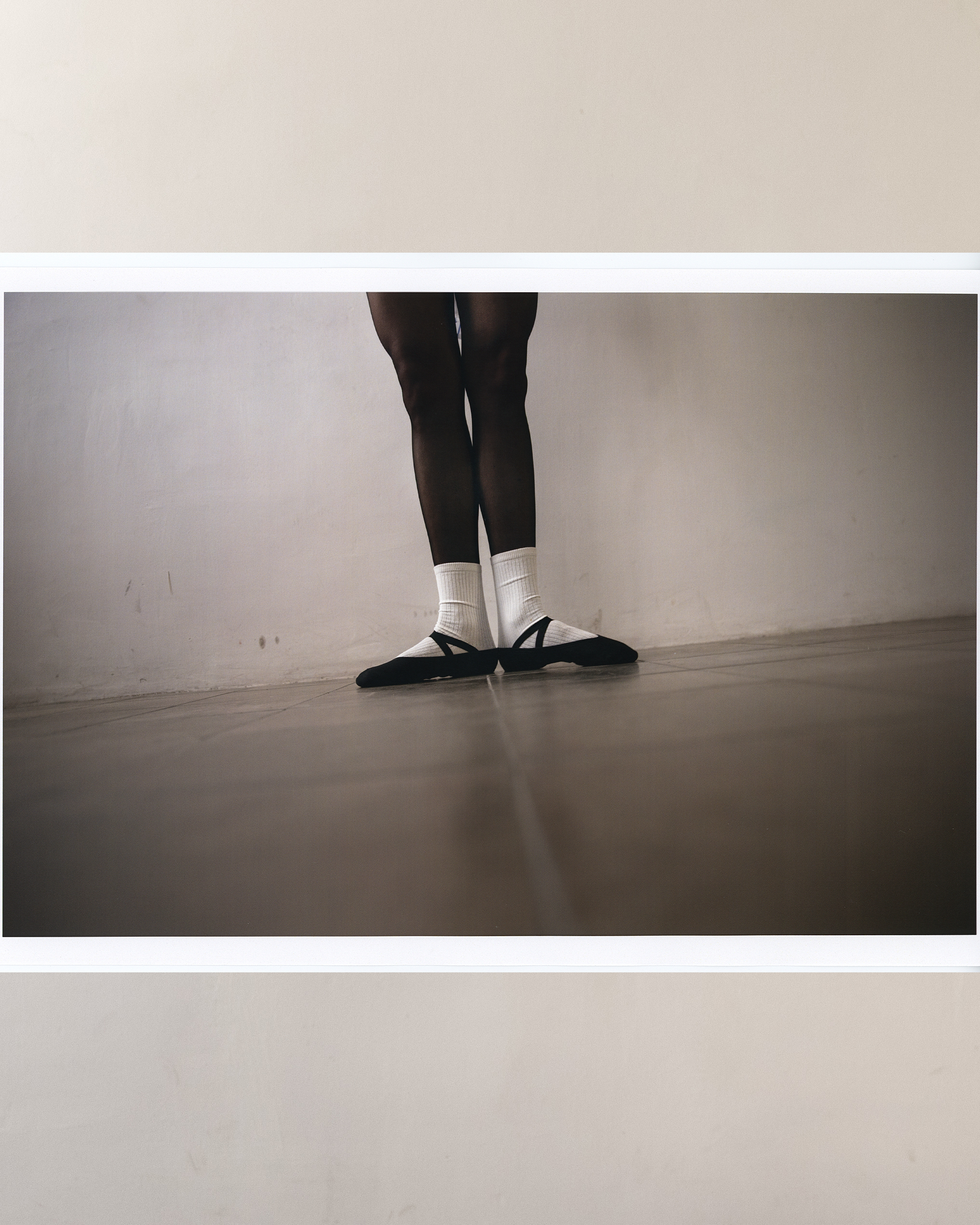 Elegant ballet dancer in black ballet shoes and white socks stands gracefully in a minimalist setting with a light wall and grey floor.