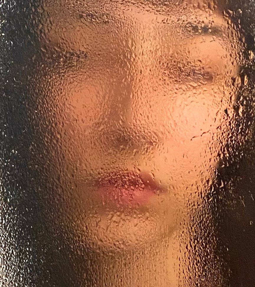 Close-up of a woman's face obscured by a surface covered in water droplets, creating a soft and diffused effect with warm lighting.