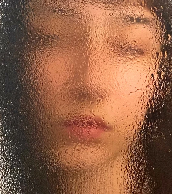 Close-up of a woman's face obscured by a surface covered in water droplets, creating a soft and diffused effect with warm lighting.