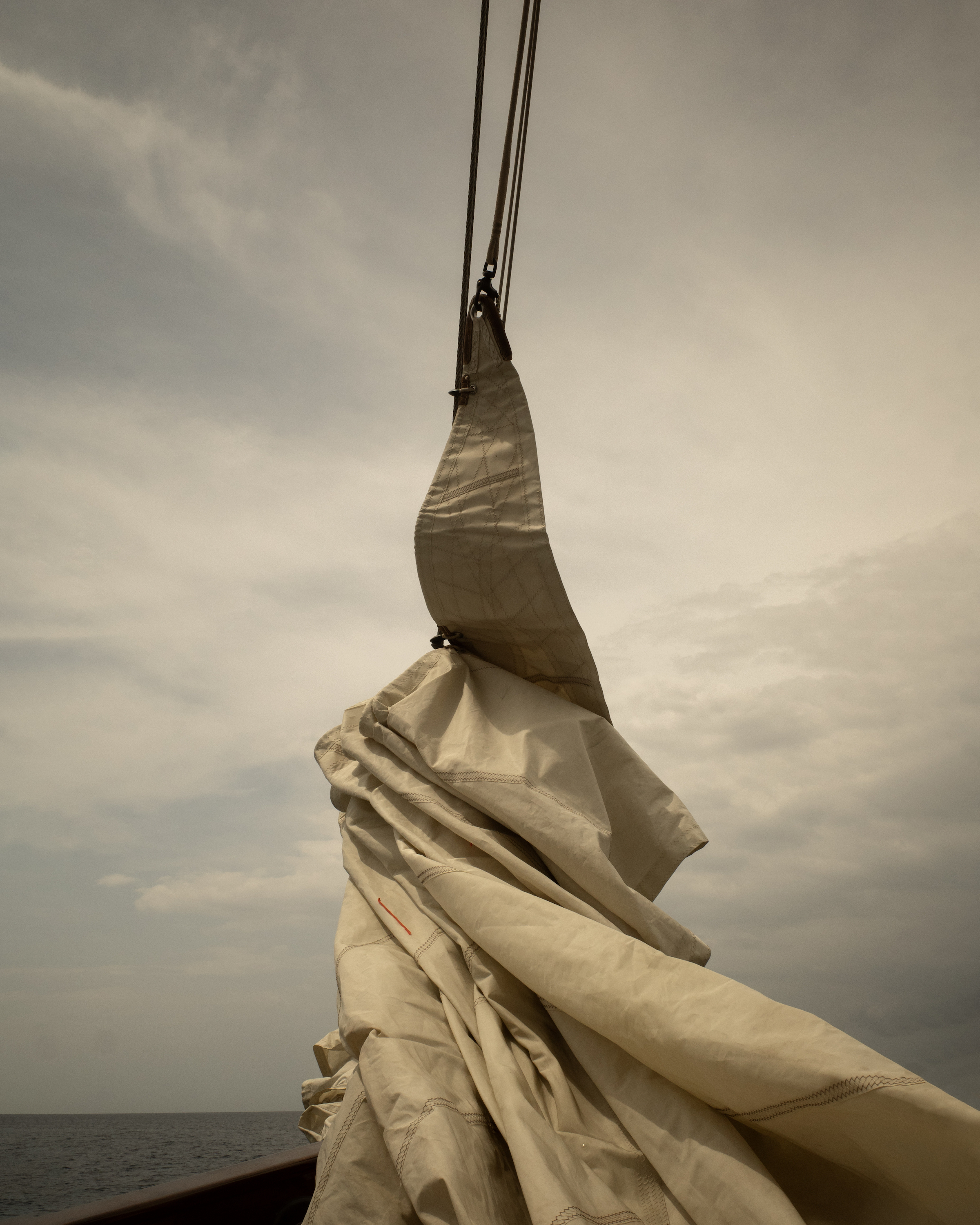 Sailboat sail partially furled with nautical ropes, ocean backdrop, overcast sky.
