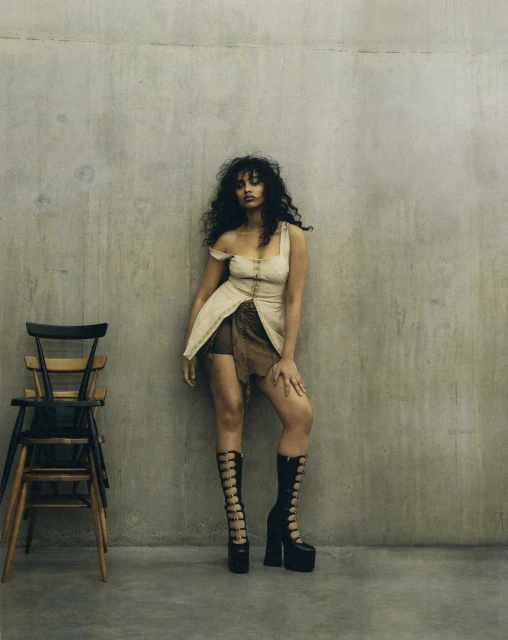 Fashionable woman with curly hair poses in a beige dress and black platform boots against a concrete wall with stacked chairs.