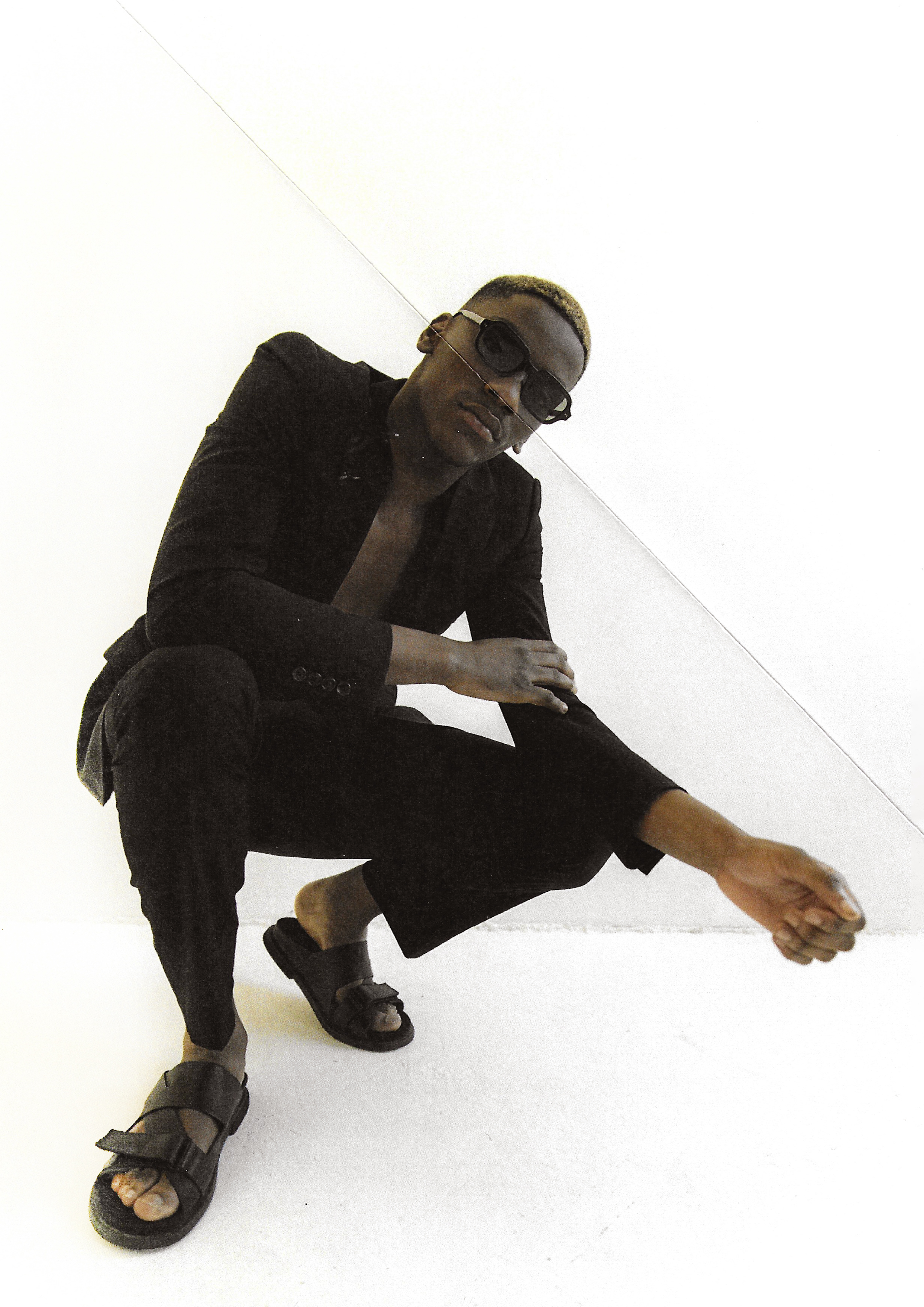 An adult Black man squats in a black suit and sandals against a white background, striking a cool pose while wearing sunglasses.