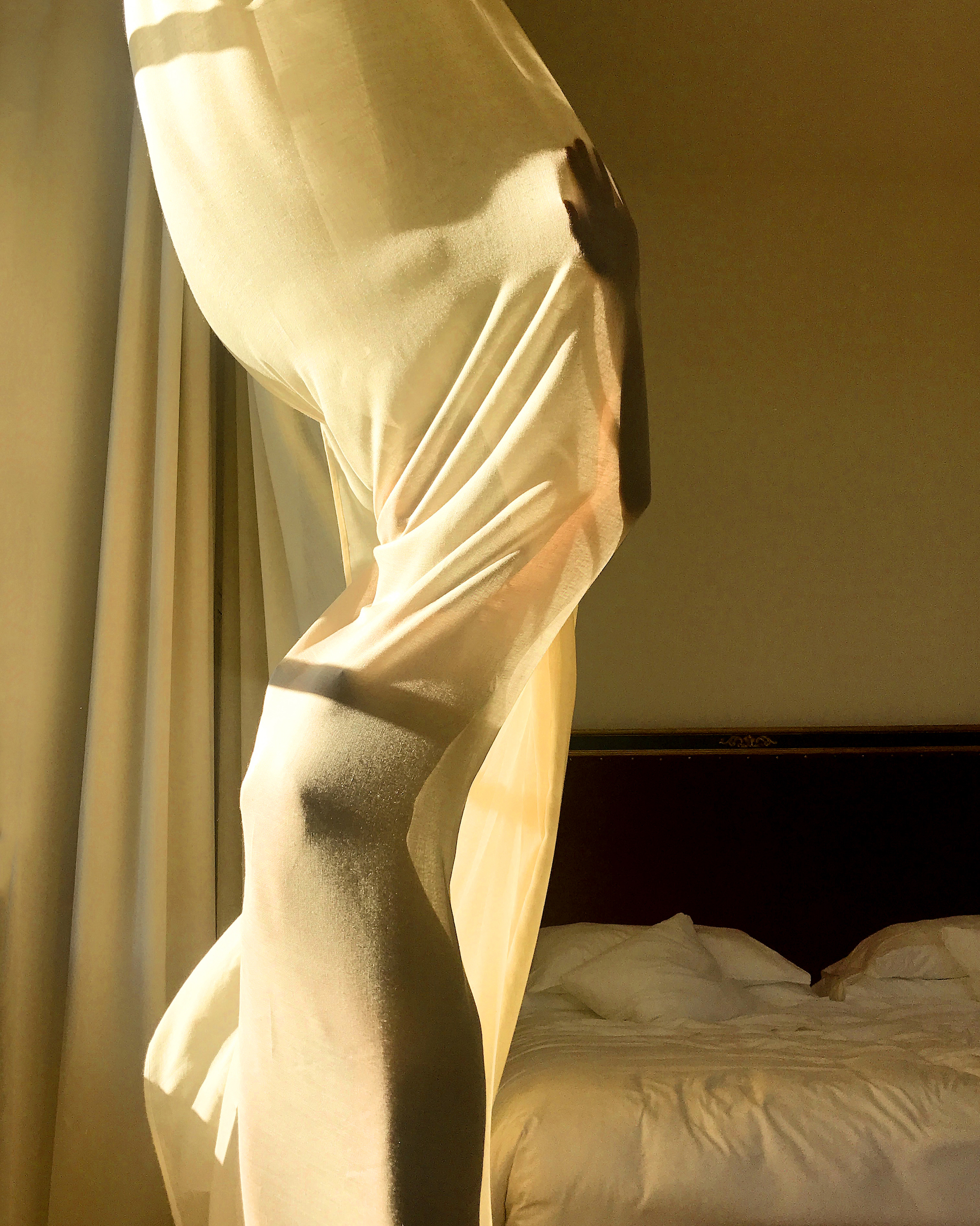 Person partially obscured by a white curtain in a warmly lit room, with a bed visible in the background, creating a soft, intimate atmosphere.