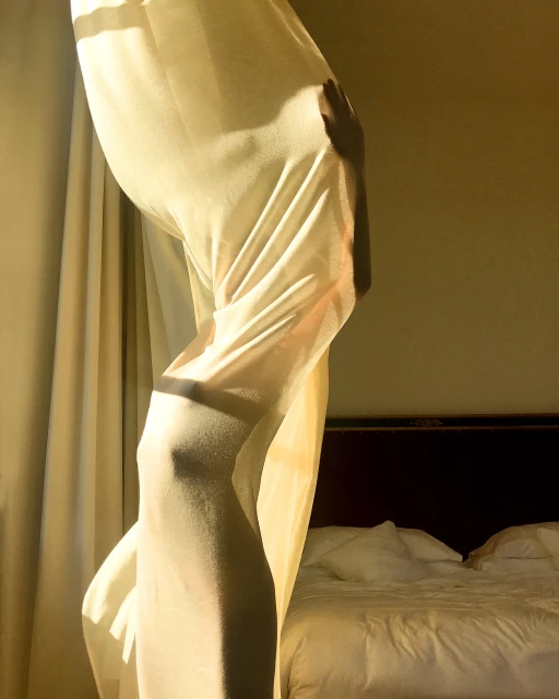 Person partially obscured by a white curtain in a warmly lit room, with a bed visible in the background, creating a soft, intimate atmosphere.