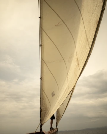 Sailboat with white sail and dark wood frame, person standing at the front, against a cloudy, overcast sky and dark water.