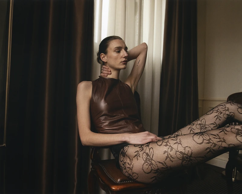 A woman in a brown leather top and patterned tights sits on a chair in a neutral-toned room with dark brown curtains and light walls.