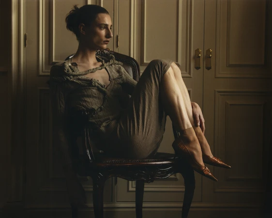 Model with hair tied up seated in a wooden chair, wearing a textured taupe dress and brown pointed heels against a paneled wall.