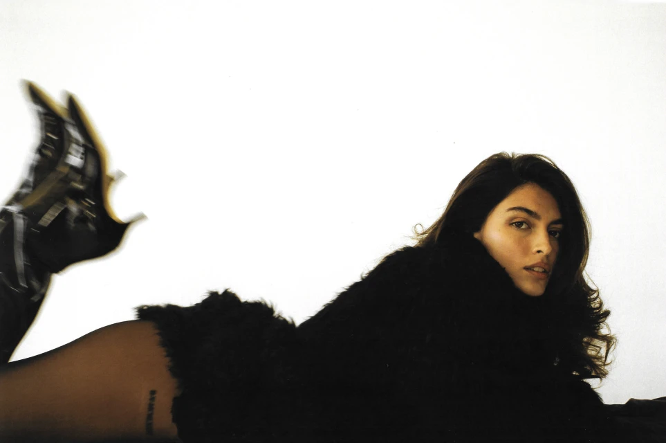 Brunette woman in a black furry jacket poses on a white background with black shiny boots blurred in motion and visible tattoos on her leg.
