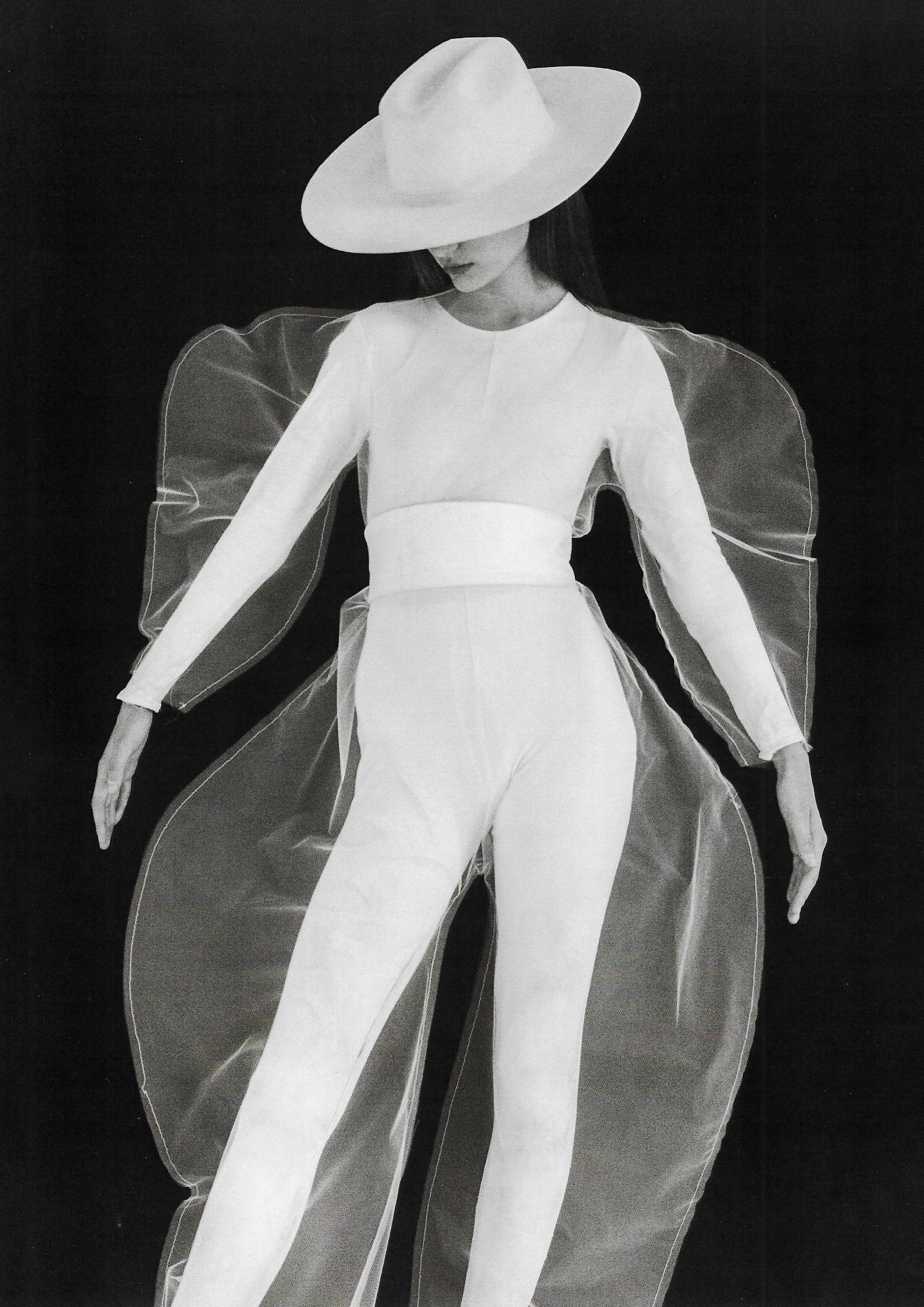 Fashion model in an all-white outfit with a wide-brimmed hat and sheer overlay against a black background in a studio portrait.