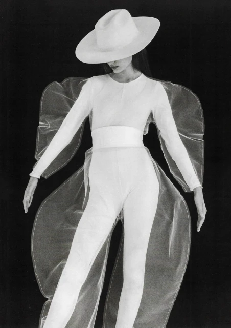 Fashion model in an all-white outfit with a wide-brimmed hat and sheer overlay against a black background in a studio portrait.