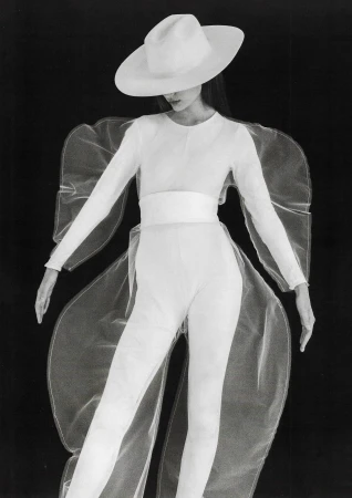 Fashion model in an all-white outfit with a wide-brimmed hat and sheer overlay against a black background in a studio portrait.
