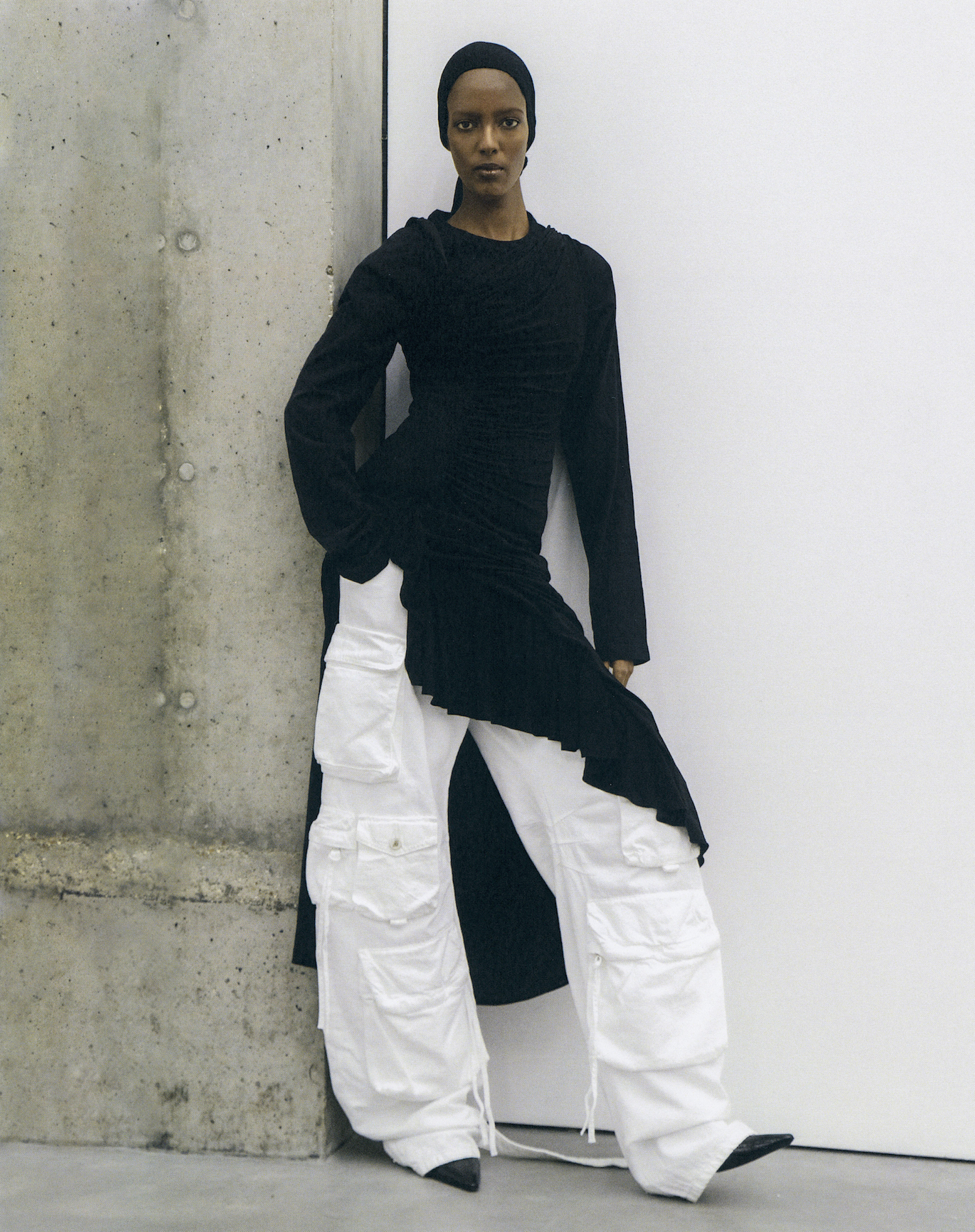 A woman with dark skin stands indoors leaning against a gray wall, wearing a black top and white cargo pants in a studio setting.