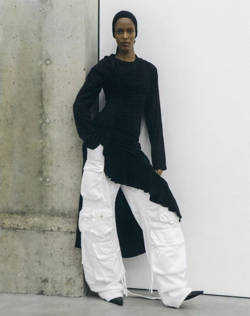 A woman with dark skin stands indoors leaning against a gray wall, wearing a black top and white cargo pants in a studio setting.