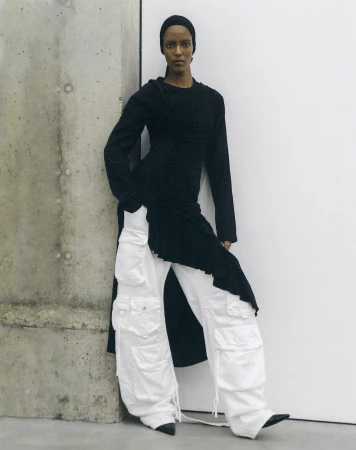 A woman with dark skin stands indoors leaning against a gray wall, wearing a black top and white cargo pants in a studio setting.