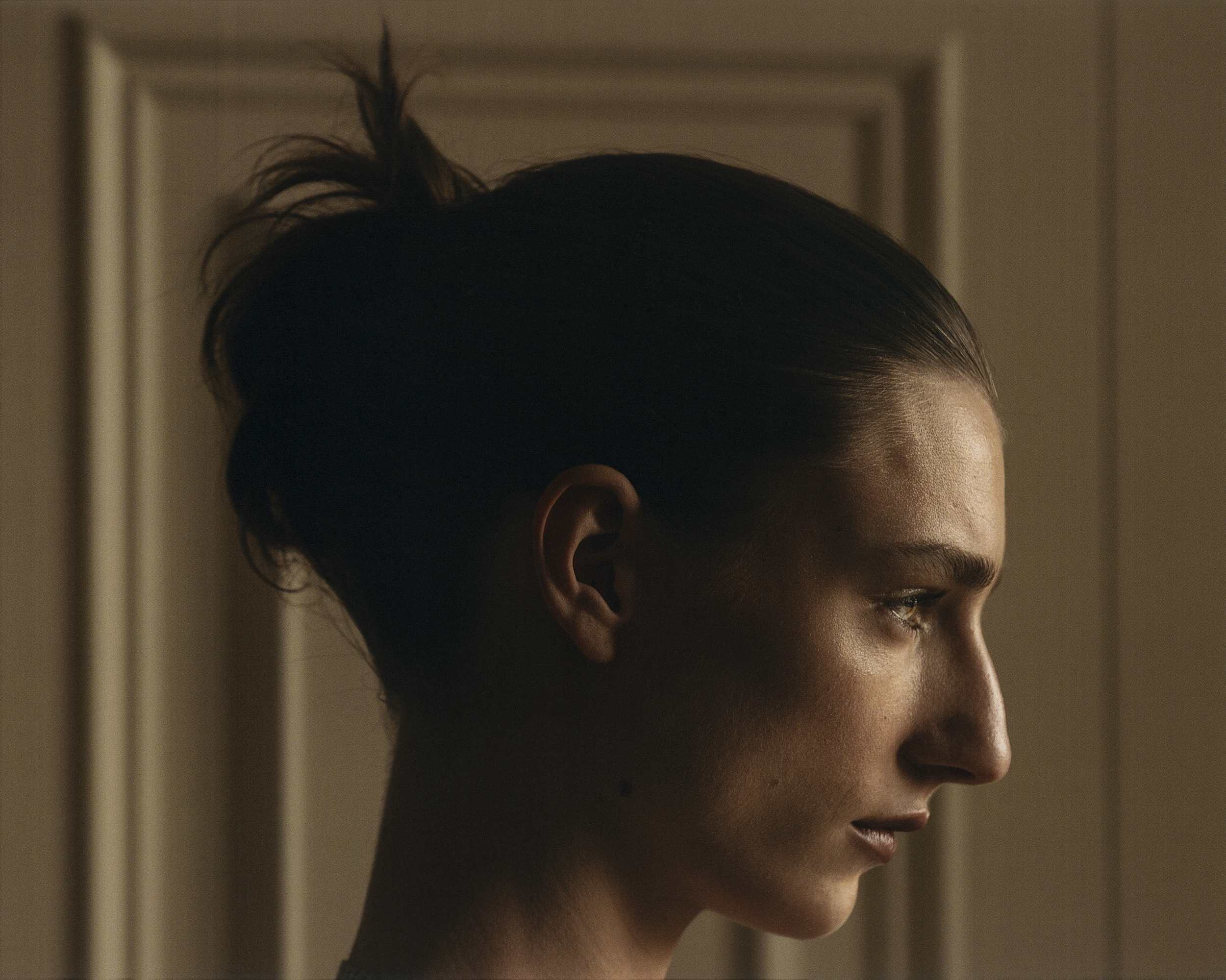 Profile portrait of a woman with dark hair tied in a bun, captured in soft lighting against a panelled wall.