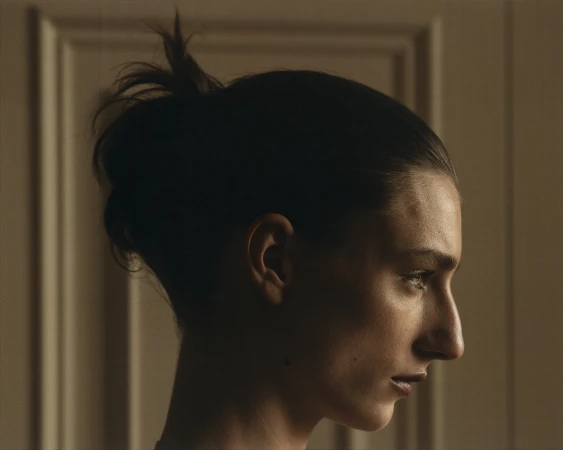 Profile portrait of a woman with dark hair tied in a bun, captured in soft lighting against a panelled wall.