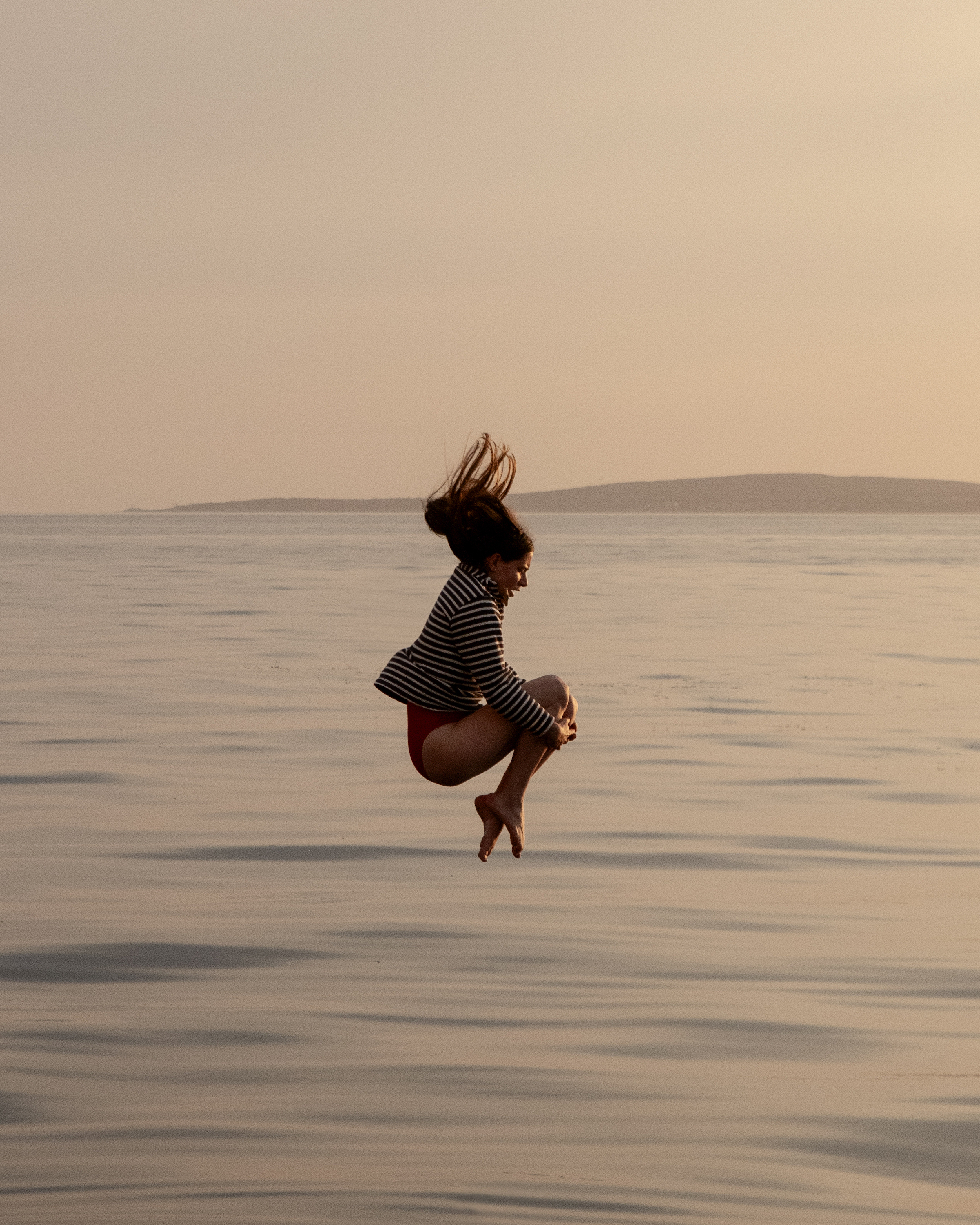 Woman in a striped blazer leaping above calm sea waters with an island visible on horizon, during golden hour with warm, neutral tones.