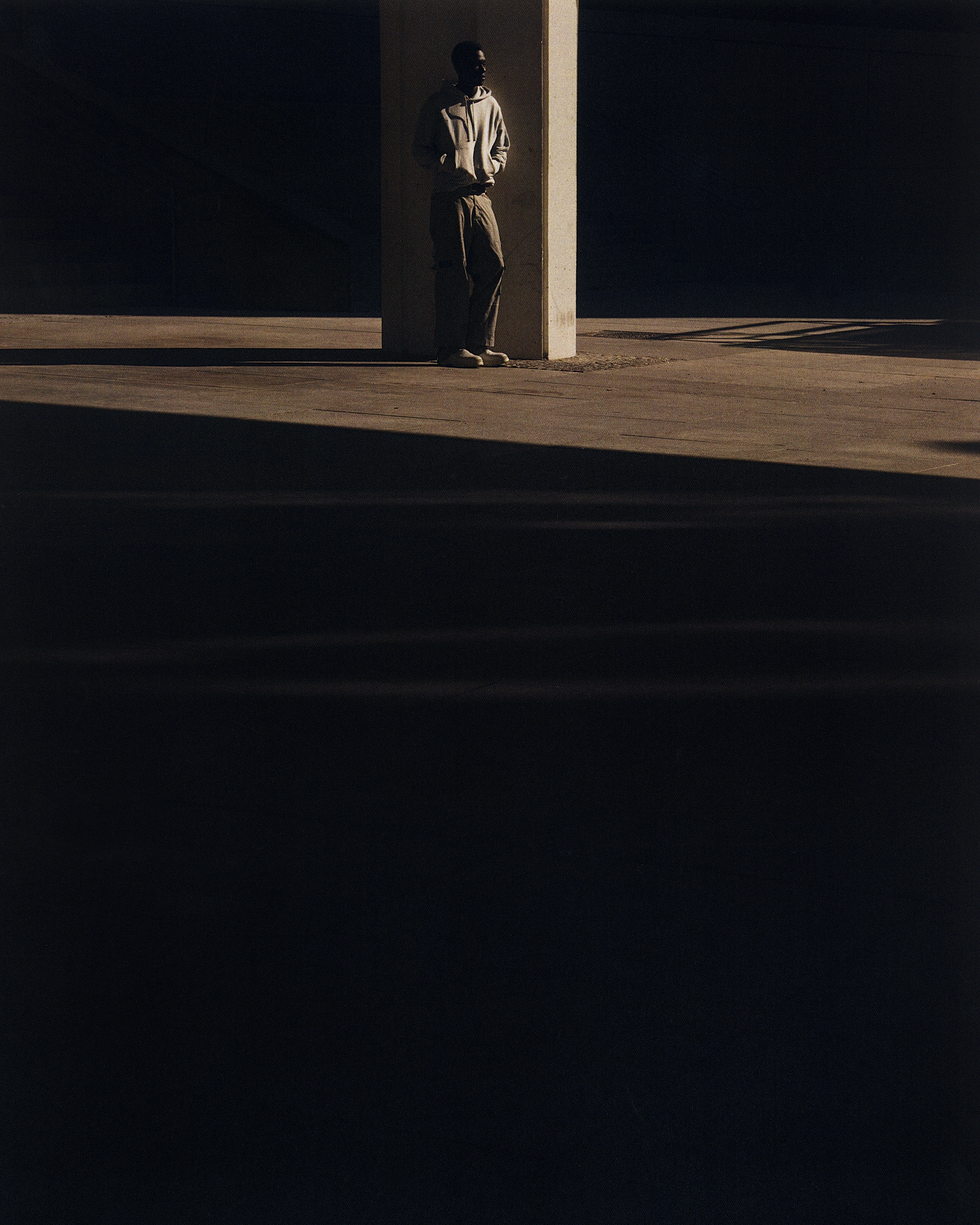 Adult male stands next to a concrete pillar in an urban environment with dark shadows and warm light.