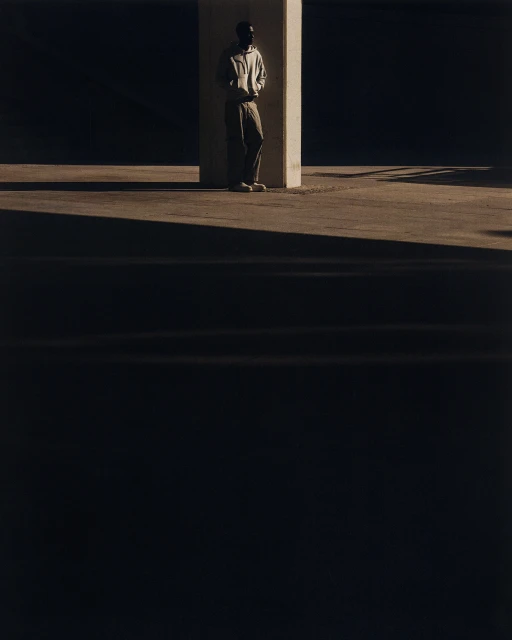 Adult male stands next to a concrete pillar in an urban environment with dark shadows and warm light.