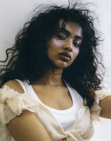 Close-up portrait of a woman with dark curly hair wearing a white tank top and ruffled blouse against a plain background.