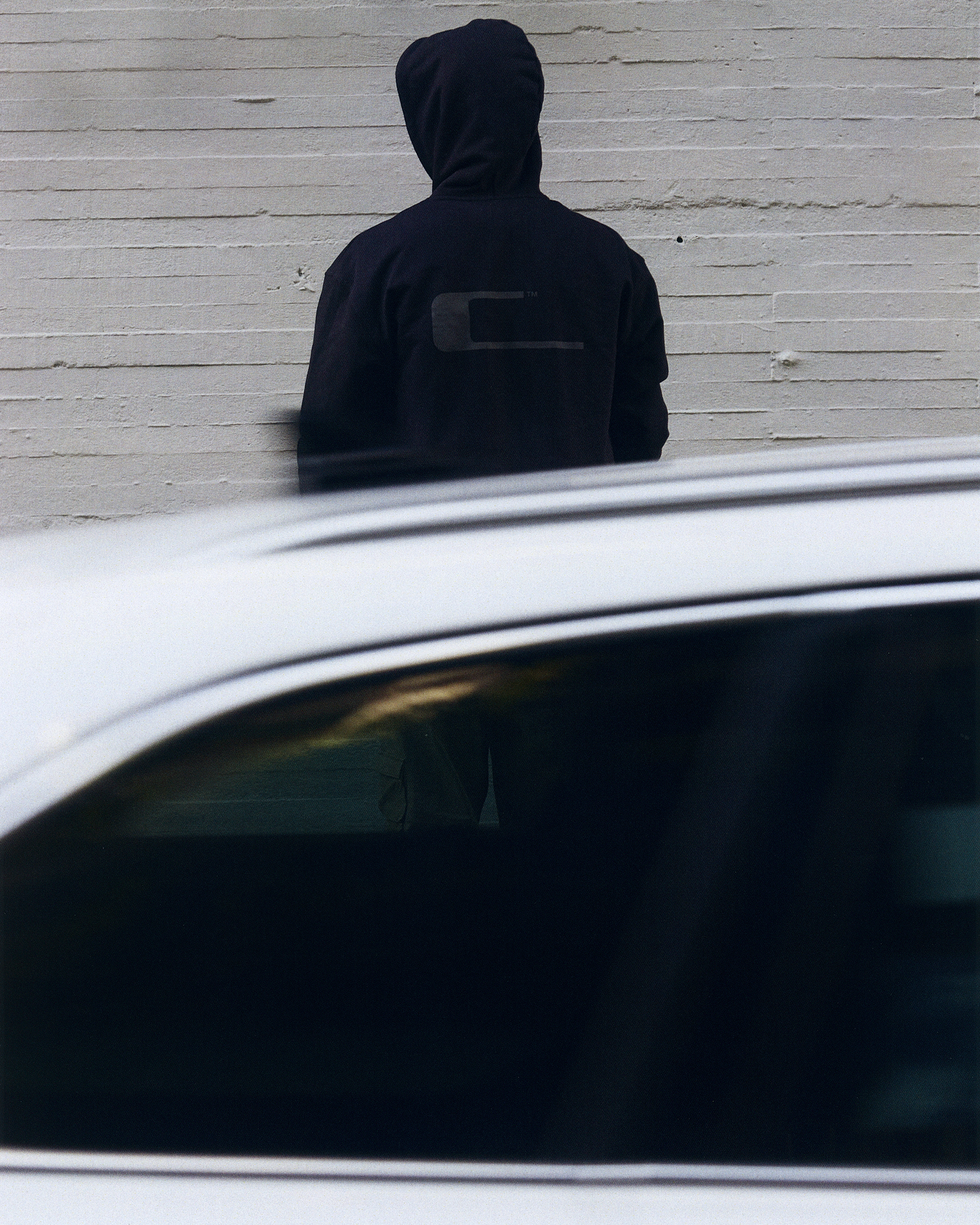 An adult is standing against a white wall with a car passing by in motion, creating a blurred effect in front of the hooded figure.