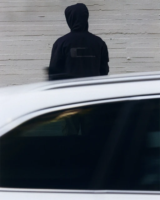 An adult is standing against a white wall with a car passing by in motion, creating a blurred effect in front of the hooded figure.