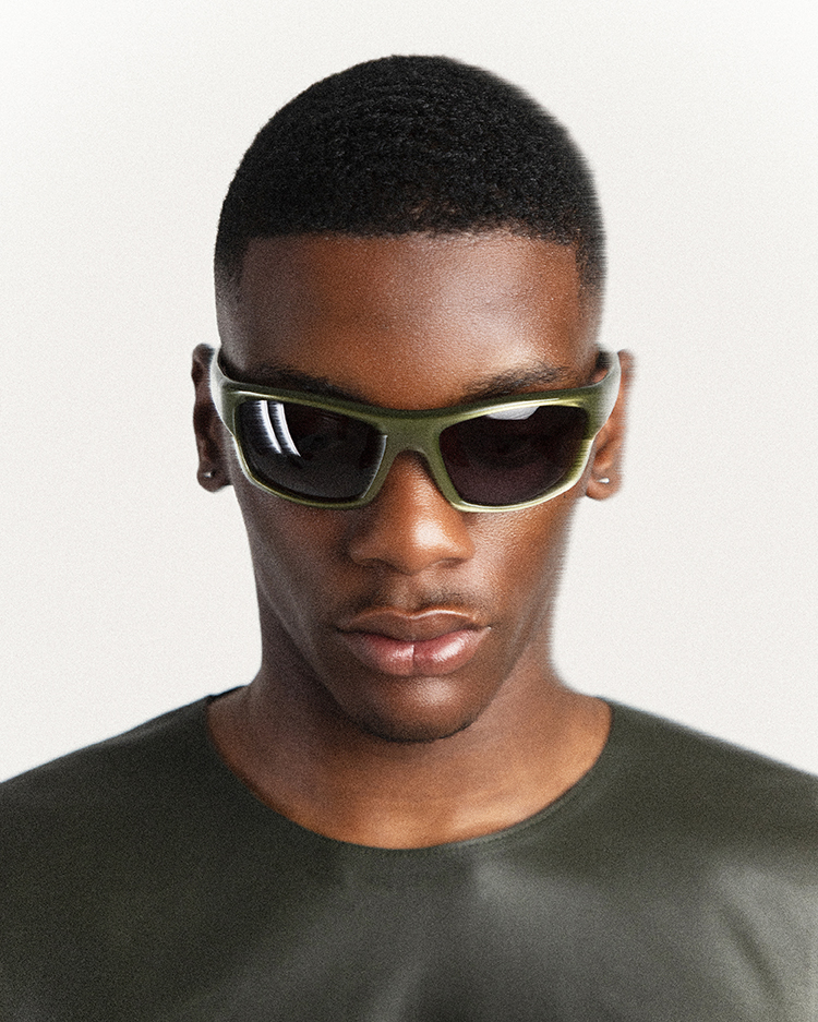 Close-up studio shot of a young Black male model wearing olive-green sunglasses and a matching shirt, looking directly at the camera.