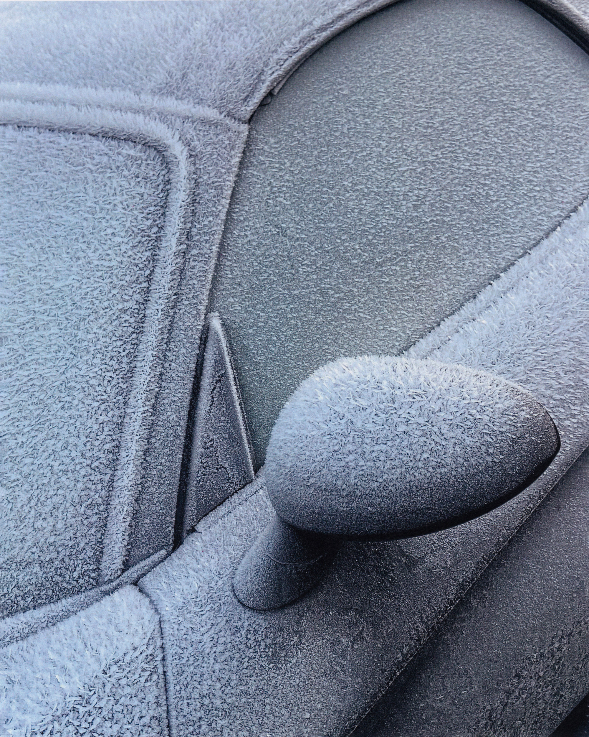 Close-up of a car covered in frost, showcasing intricate ice crystals on the windows, side mirror, and body panels in an outdoor setting.