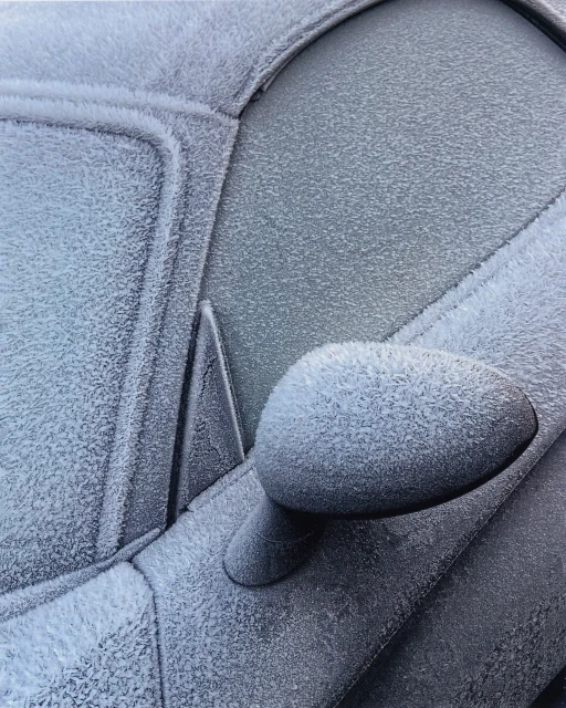 Close-up of a car covered in frost, showcasing intricate ice crystals on the windows, side mirror, and body panels in an outdoor setting.