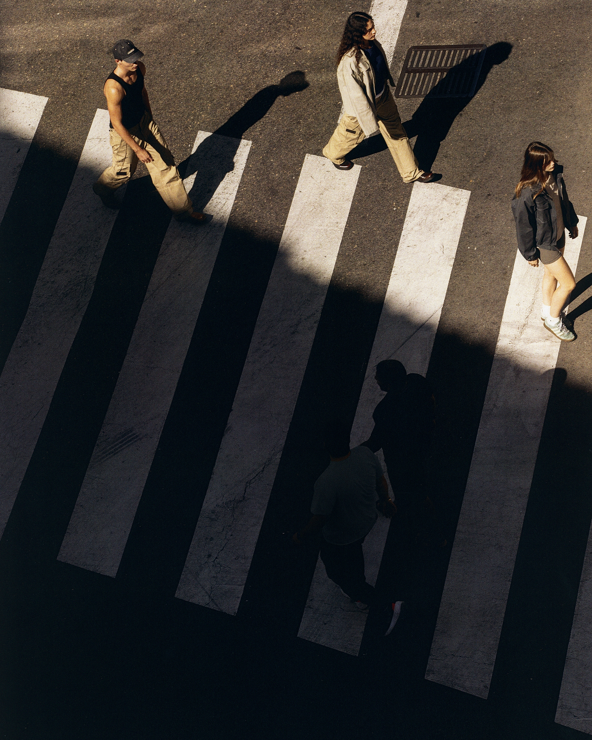 Aerial view of three people in casual outfits crossing a pedestrian crosswalk with zebra stripes on an asphalt road on a sunny day.
