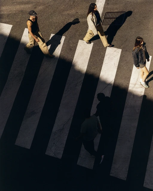 Aerial view of three people in casual outfits crossing a pedestrian crosswalk with zebra stripes on an asphalt road on a sunny day.