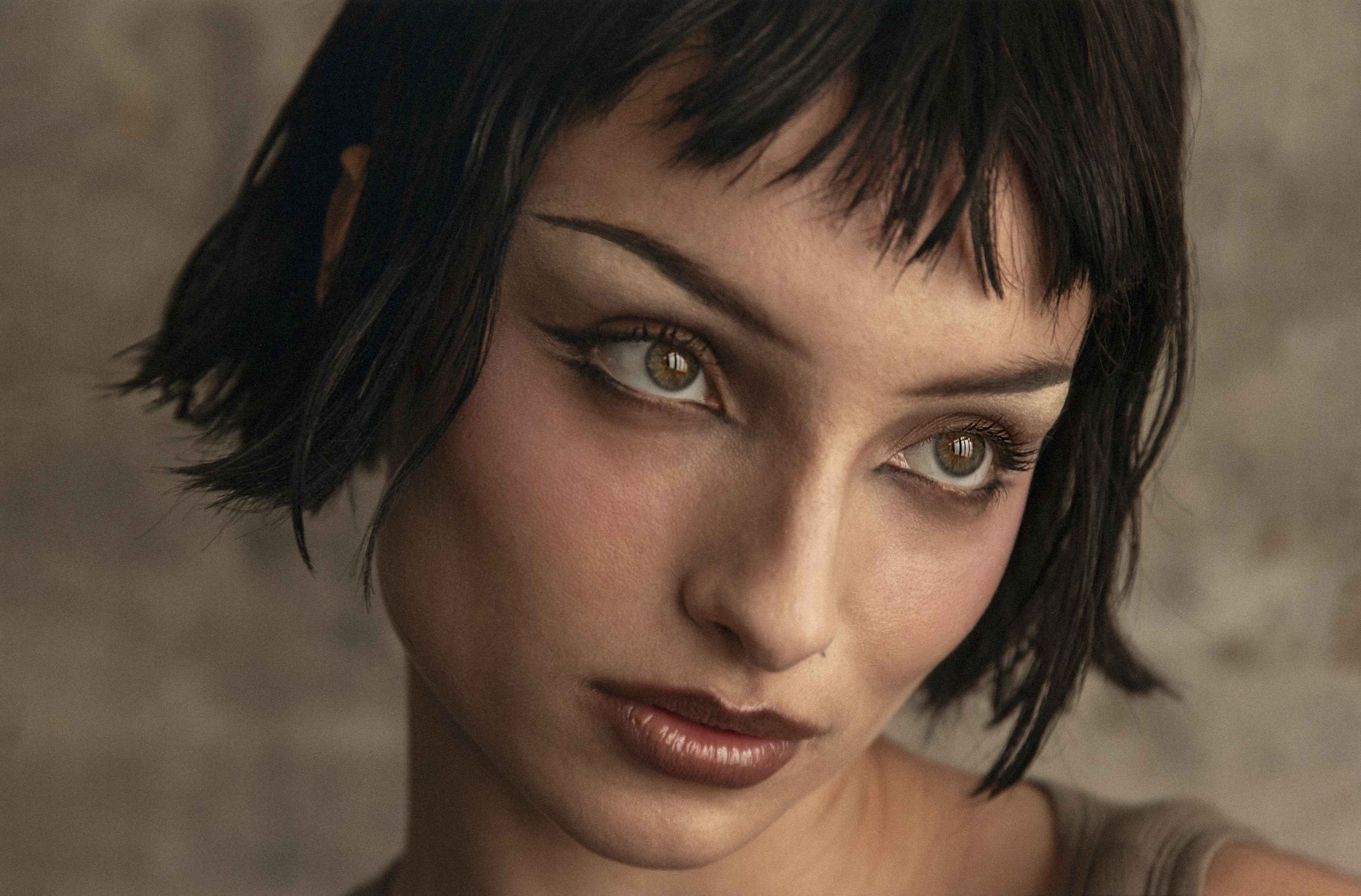 Close-up portrait of an adult woman with short black hair, dramatic eye makeup, and glossy brown lips, posing in front of a textured background.