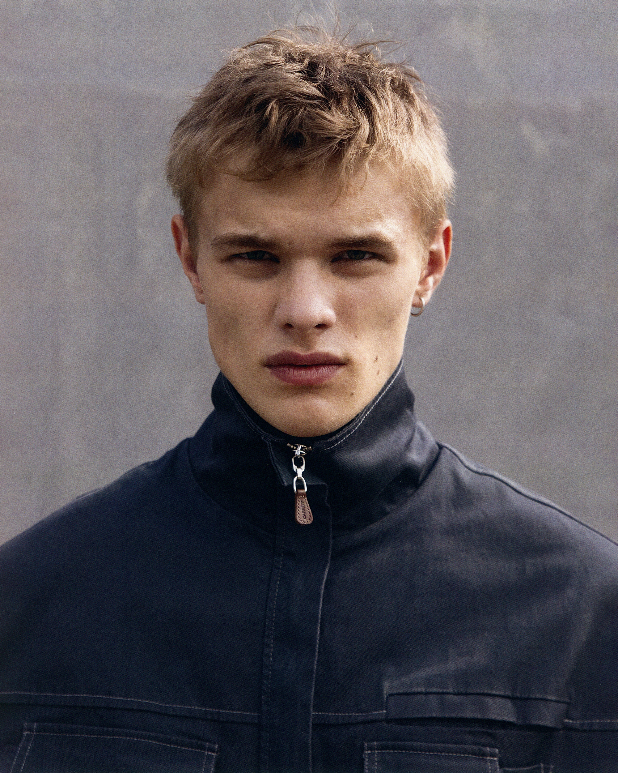 Close-up portrait of a blonde male model wearing a black jacket, set against a neutral grey background in a studio setting.