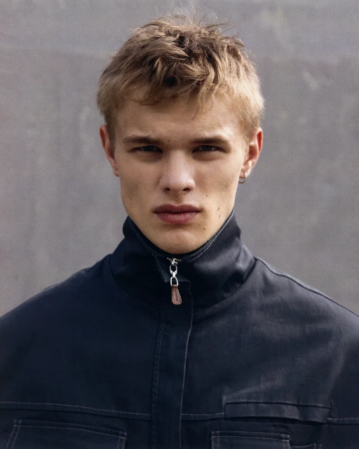 Close-up portrait of a blonde male model wearing a black jacket, set against a neutral grey background in a studio setting.