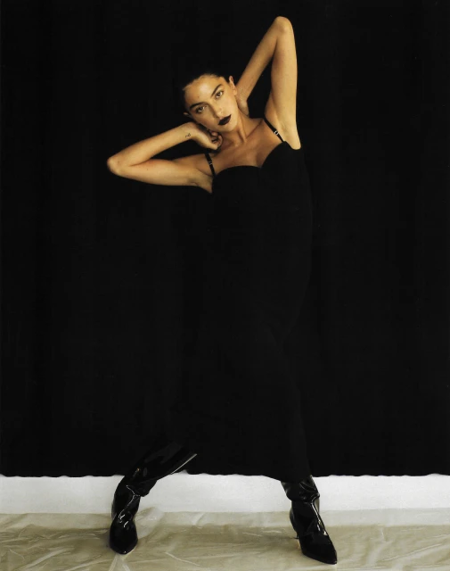 A slender woman in a black dress and shiny black boots poses against a dark backdrop in a studio shot, arms raised to her head.