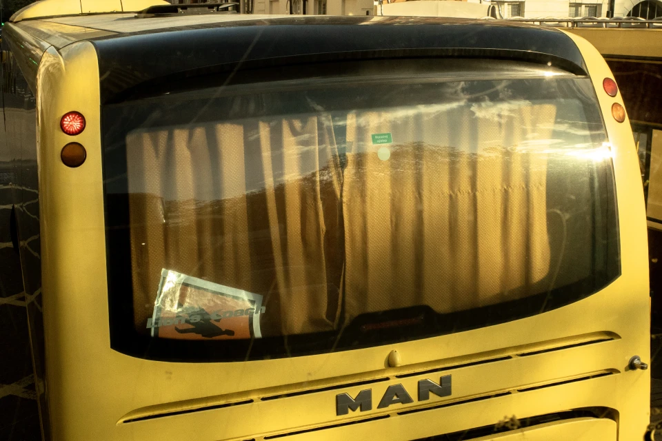 Rear view of a yellow bus with curtains in the back window and the MAN logo visible, parked outdoors on a sunny day.