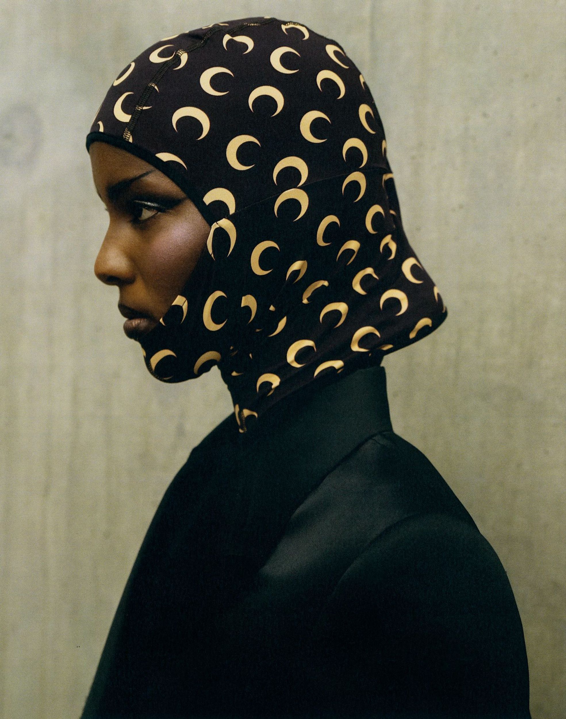 Profile shot of a dark-skinned model wearing a black jacket and head covering patterned with gold crescents against a grey wall.