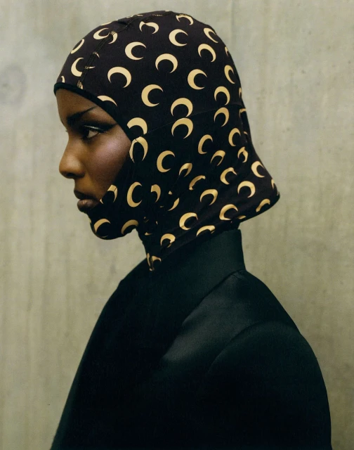 Profile shot of a dark-skinned model wearing a black jacket and head covering patterned with gold crescents against a grey wall.