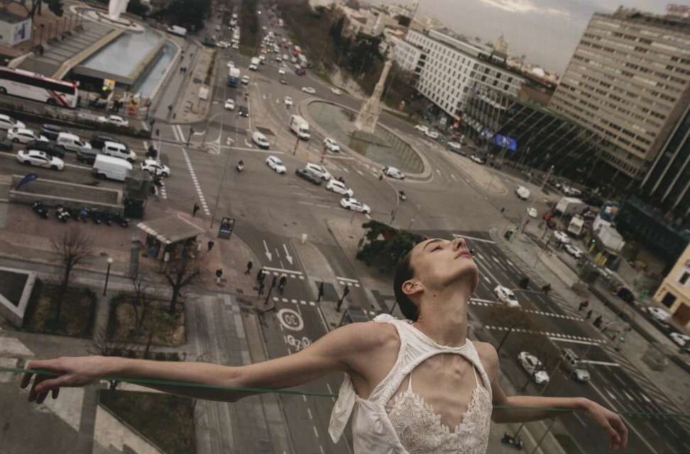 High-angle view of an adult female model in a white dress on a rooftop with a cityscape background featuring busy streets and buildings.