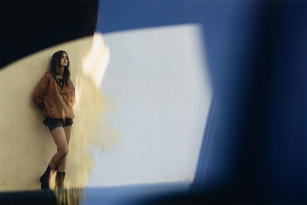 A young woman leans against a cream-colored wall in an outdoor setting, illuminated by a soft light and framed by shadowed edges.
