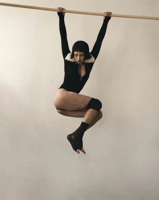 Athletic woman in a black bodysuit and socks hangs from a wooden bar in a neutral studio, showcasing strength and body flexibility.